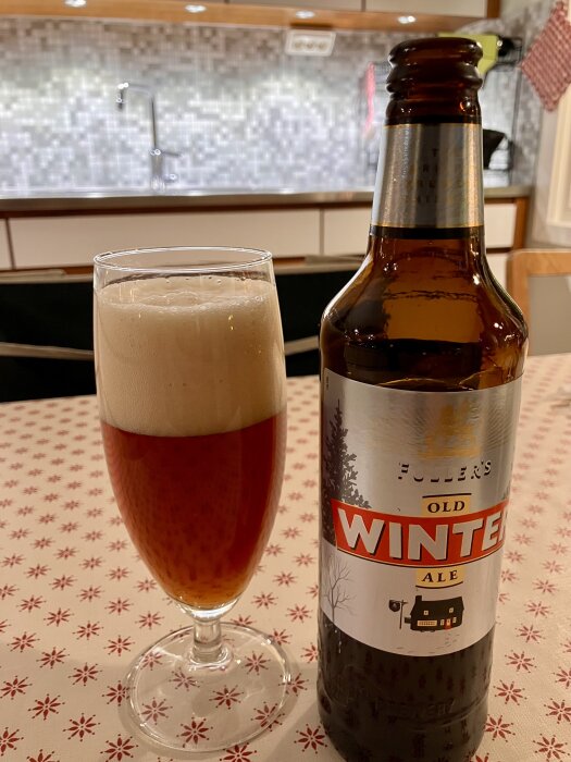 Ett glas med skummande öl bredvid en flaska Fuller's Old Winter Ale på ett bord med stjärnmönstrad duk och kök i bakgrunden.