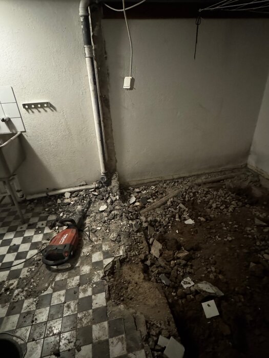 Rivningsarbete på källargolv med synligt fundament av betong och en borrhammare. Rester av kakel och jord ligger på golvet. Vägg med rör syns i bakgrunden.