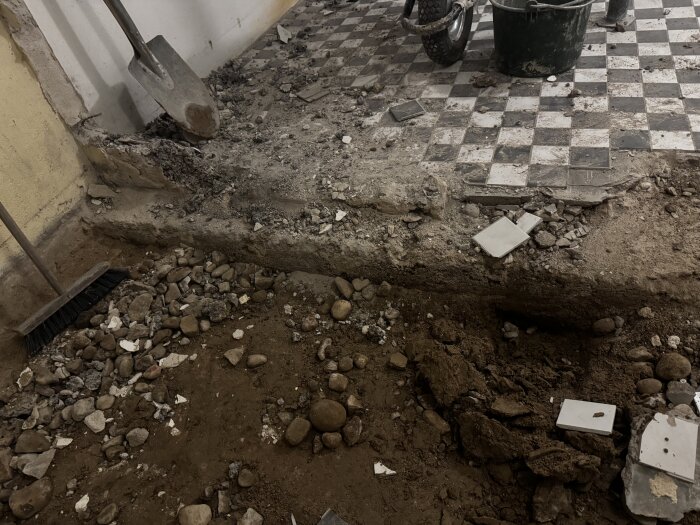 En källare med pågående golvarbete, synligt betongfundament, lös jord och stenar, kakelbitar och verktyg som en spade och borste.
