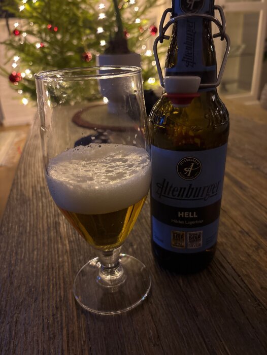 Glas med skummande öl och en flaska med etiketten "Altenburger Hell Mildes Lagerbier" på ett träbord, med en ljusprydd julgran i bakgrunden.