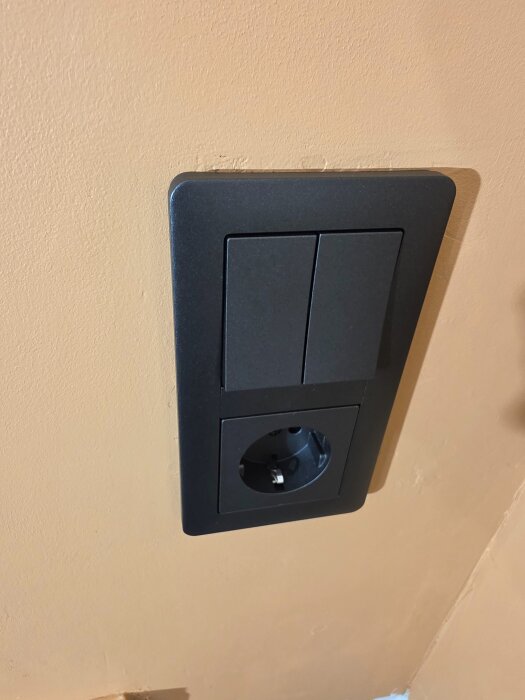 Svart vägguttag med två strömbrytare monterat på en beige vägg.