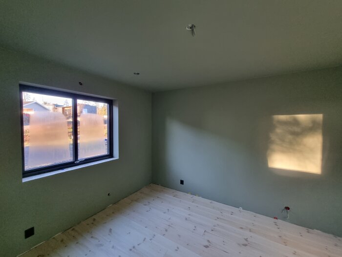Ljust rum med nylagt ljust trägolv och stort fönster med utsikt mot hus; solbelyst vägg.