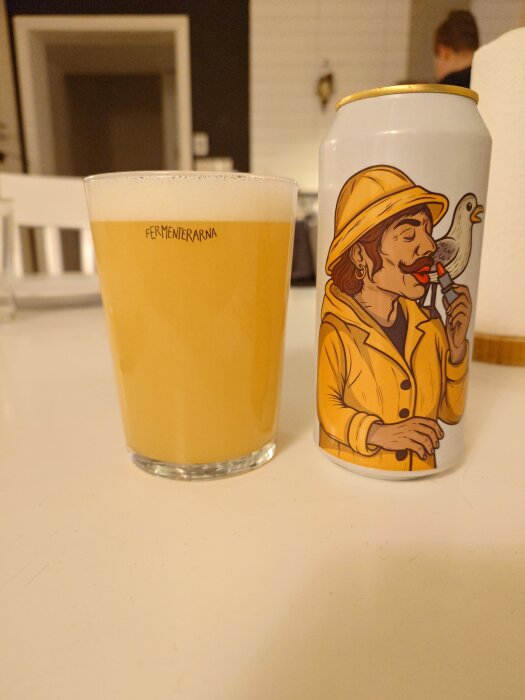 Ett glas öl och en burk med illustration av en person i gul jacka och hatt med en fågel. Märket är Fermenterarna.