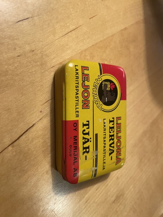 Gul och röd plåtburk för Leijona lakritspastiller med text "LEIJONA TERVA-TJÄR-" och tillverkad av Oy Merijal AB, ligger på ett träbord.