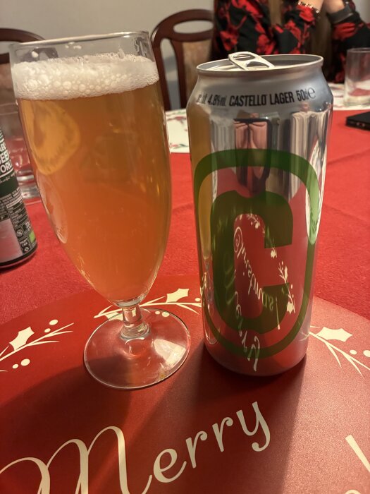 Ett glas öl bredvid en burk Castello Lager på ett rött bord med julig dekoration och texten "Merry".