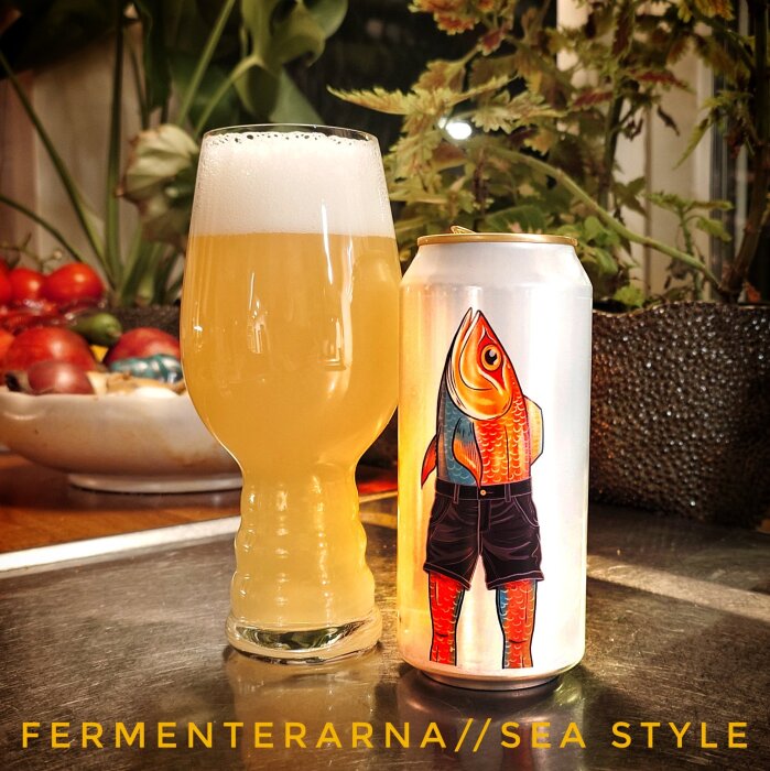 Ett glas ljus öl bredvid en burk med färgglad fiskillustration, omgivna av växter och tomater, med texten "FERMENTERARNA//SEA STYLE".