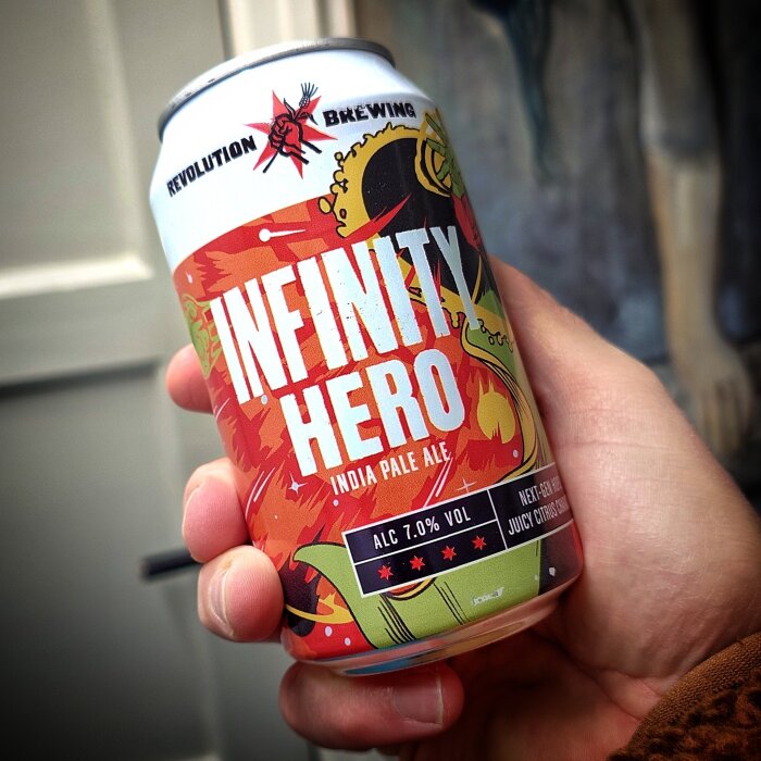 En hand håller en burk Infinity Hero India Pale Ale från Revolution Brewing med färgglad etikett.