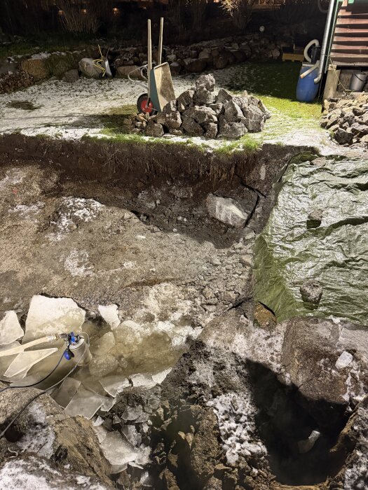 Byggarbetsplats med jordgrop, stenar och en skottkärra. Isbitar och vattenpump i gropen. Bakgrunden visar gräsmatta täckt av snö.