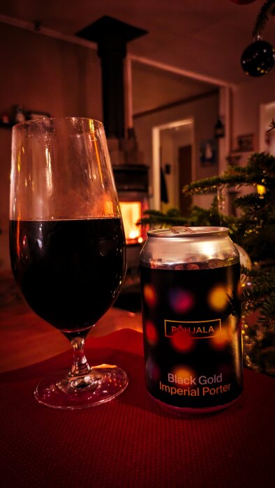 Glas och burk med Põhjala Black Gold Imperial Porter framför en tänd kamin och en julgran, i mysig kvällsbelysning.