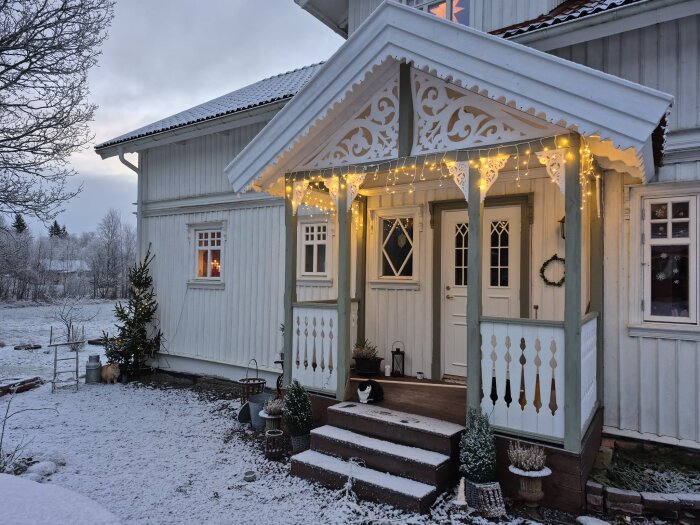 Frostig trädgård och veranda dekorerad med julgransljus på ett vitt hus, snötäckt mark och vinterlandskap i bakgrunden.