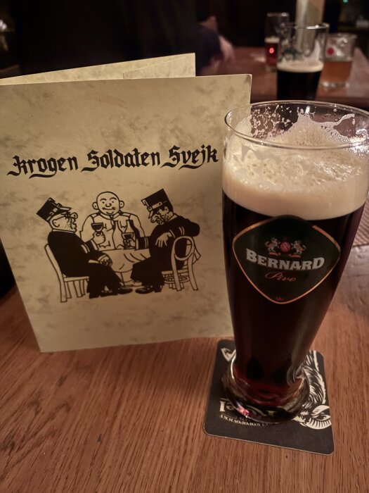 Ett glas öl från Bernard står på ett bord bredvid en meny från Krogen Soldaten Svejk. I bakgrunden syns fler ölglas på ett bord.