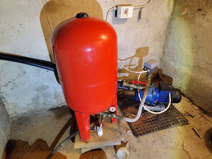 En röd hydropress med tillhörande blå vattenpump sitter på en betongvägg. Rör och ventiler kopplas till pumpen.