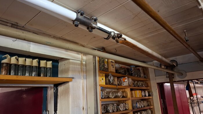Rörinstallation i ett hem med vita och kopparrör, monterade i taket ovanför hyllor med burkar och skruvförvaring.