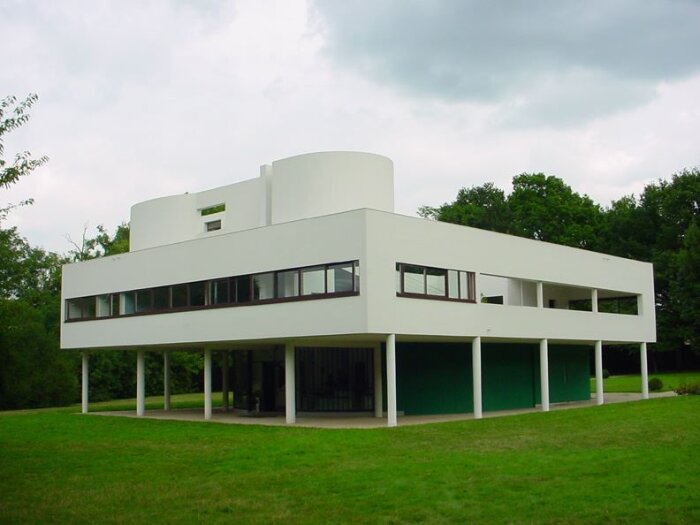 Modernistiskt hus med vita väggar och långa fönster, placerat på pelare med grönt gräs runtomkring.