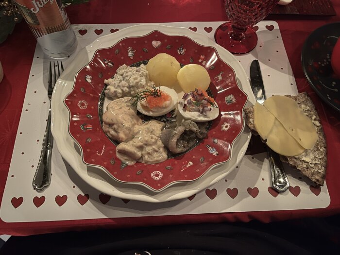 Tallrik med julmat: potatis, sill, ägg med rom och dill på röd tallrik, bröd med ost bredvid, på julduk med rött vinglas och julöl i bakgrunden.