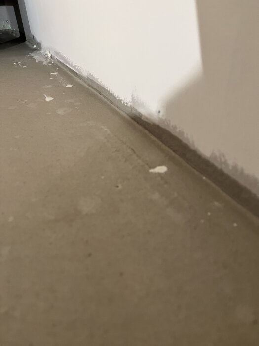 Upphöjt glasfibernät syns under flytspacklat golv nära vägg. Små trämarkörer och oregelbunden yta indikerar potentiella problem vid golvläggning.