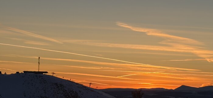 Solnedgång över en bergstopp med skidliftar och snö, omgiven av färgade molnränder på himlen.