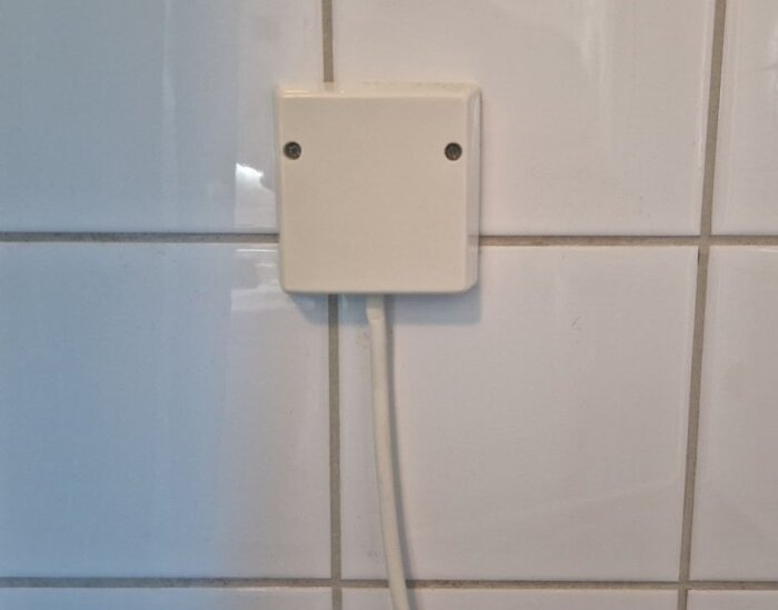 En vit kopplingsdosa monterad på kakelvägg, med sladd nedåt.