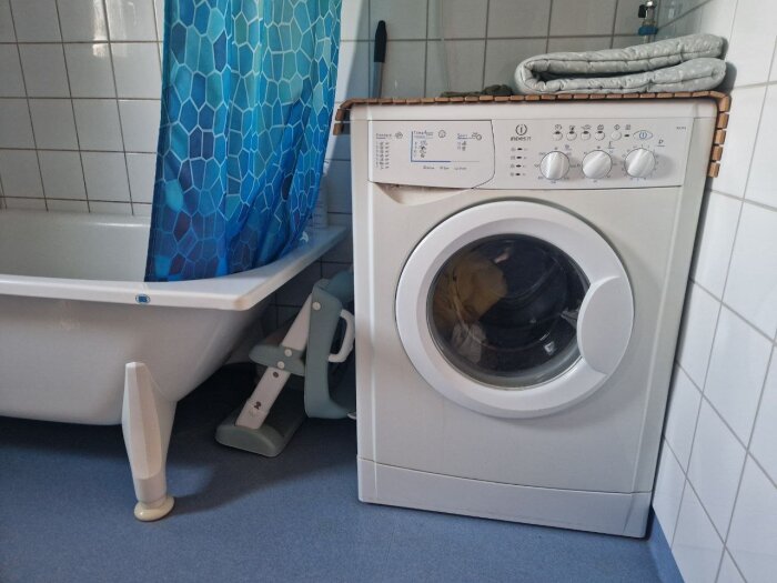 Tvättmaskin nära badkar i ett badrum, med en blå duschdraperi och grå badrumsmatta.