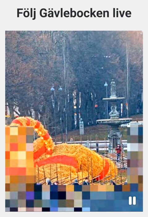 Halmget dekorerad med rött band under konstruktion, omgiven av stängsel och arbetare. Bakgrund med träd och en fontän.