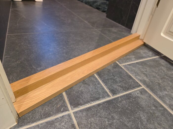 Platsbyggd tröskel av träbitar på ett mörkt klinkergolv mellan två rum.