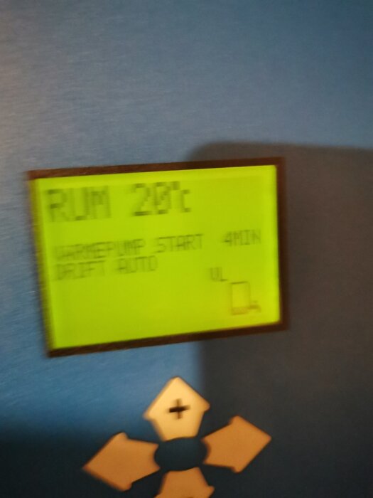 Display på värmepump som visar "Rum 20°C", "Värmepump start 4 min", med pilar och en plusknapp under.