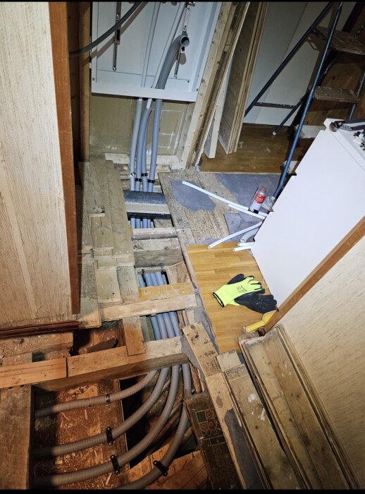 Golv under renovering med öppet träbjälklag som blottar grå rörinstallationer. En hylla, handskar och verktyg syns intill.
