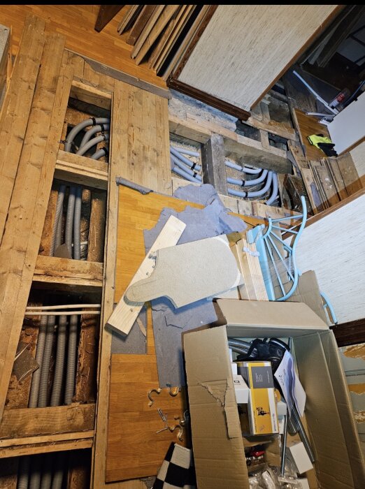 Golv i renovering med synliga rör och isolering. Verktyg och byggmaterial, inklusive kartong och blå rör, ligger på golvet.