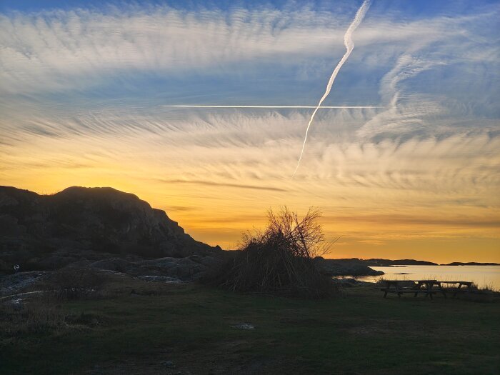 Solnedgång vid havet med flygspår på himlen, klippiga kullar och en picknickbänk i förgrunden.