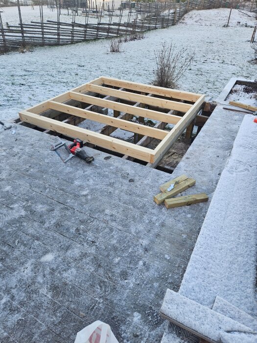 Byggarbetsplats med trästomme för en byggnad, omgiven av snötäckt mark och staket i bakgrunden. Verktyg och träbitar ligger på marken.