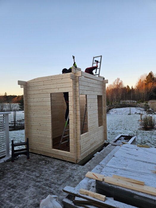 Byggprojekt i trä, två personer arbetar på taket av en liten konstruktion i kallt väder med snö på marken.