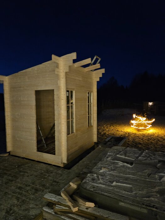 En halvfärdig trälada under uppförande på kvällen, omgiven av snö och byggmaterial. I bakgrunden brinnande eld.