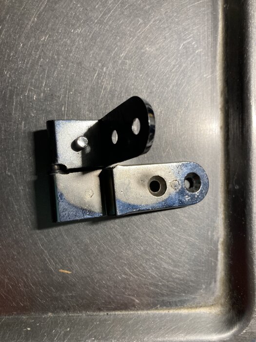 Gångjärn i metall med flera hål för montering, troligen för skåpdörrar eller fönster, mot en bakgrund av en metallisk yta.