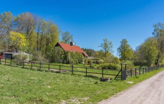 Rött hus med tegeltak i lantlig miljö, omgiven av grönskande träd och en grusväg. Staket omger tomten, tydligt vårväder.