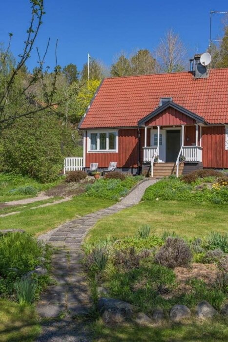 Rött hus med vita detaljer, trädgård och stenlagd gång nära skog. Kåpan är brant och taket har betongpannor.