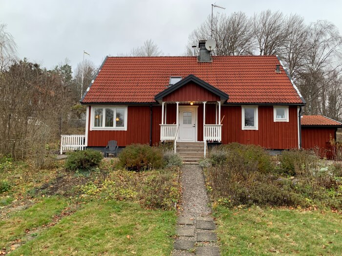 Rött hus i två våningar med vit dörr och veranda. Taket har röda tegelpannor och omgivningen präglas av buskage och vinterkala träd.