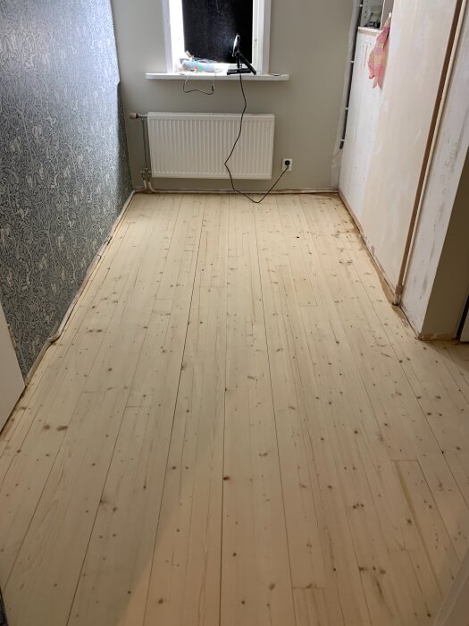 Nyslipat grangolv i sovrum med vitmålade väggar och ett mönstrat tapetparti, synlig radiator under fönstret.
