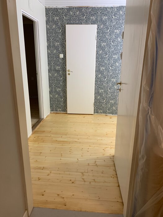 Nyrenoverad nisch med ljusa trägolv och en vit dörr, väggarna har mönstrad fondtapet.