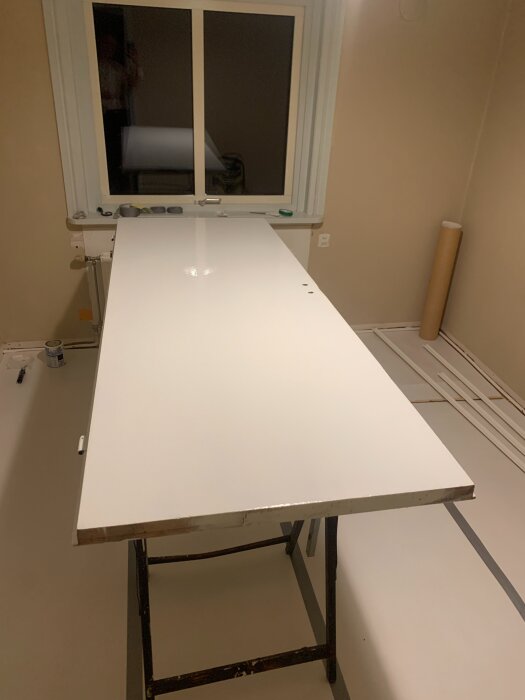 Vit dörr som står på ett par bockar för målning i ett rum med beigea väggar och vitt golv. I bakgrunden syns ett fönster.