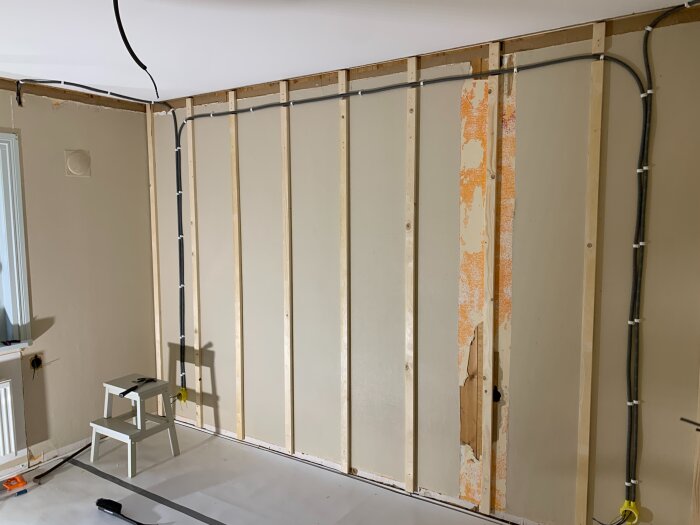 Renovering av ett rum med synliga reglar, kablar, och gammal tapet som skrapats bort. En liten stege står på golvet.