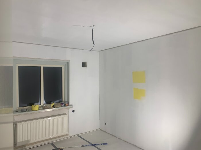 Rum under renovering med nyligen målade väggar, färgprover i gult på ena väggen, fönster och värmeelement synliga.