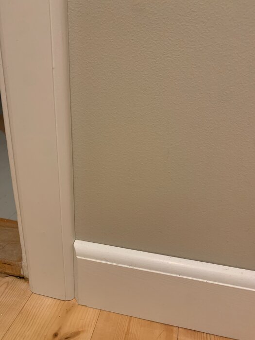 Vitt dörrfoder och golvlist på ljus träparkett och beige vägg i ett rum.