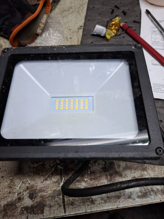 En trasig Biltema LED-armatur på ett arbetsbord med verktyg och papper runtomkring.