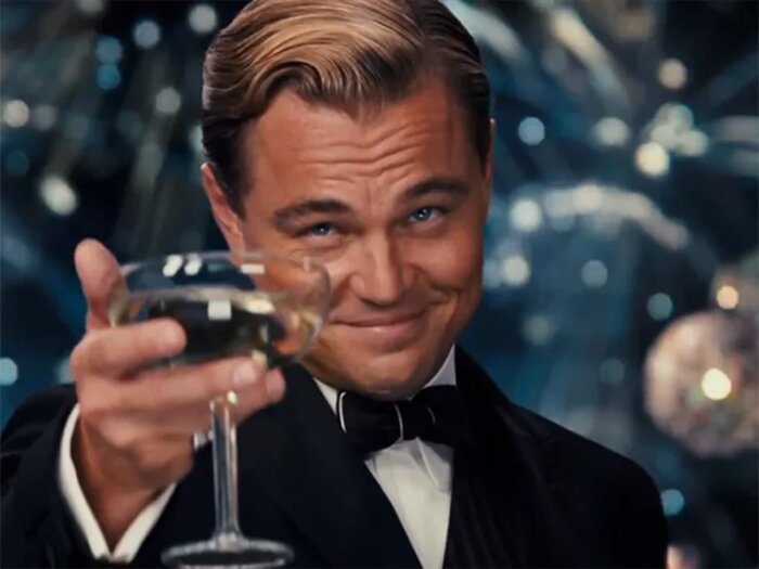 En man i smoking lyfter ett glas champagne i en festlig miljö.