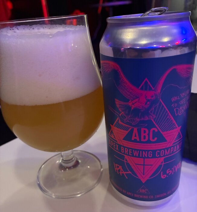 Ett glas öl bredvid en burk Apex Brewing Company IPA med en uggla och texten "ABC".