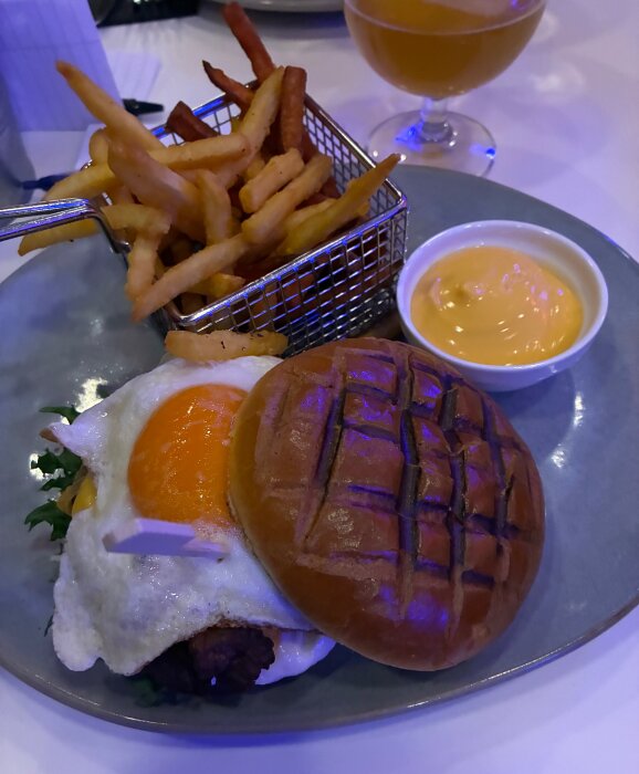 Crazy burgare med ägg och pommes frites i korg samt cheddardipp på en tallrik, med ett glas dryck i bakgrunden.