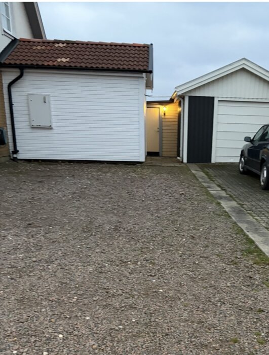 Bild av en vit byggnad med sadeltak som hänger ihop med ett hus. Till höger syns grannens förråd och en bil delvis parkerad på uppfarten.