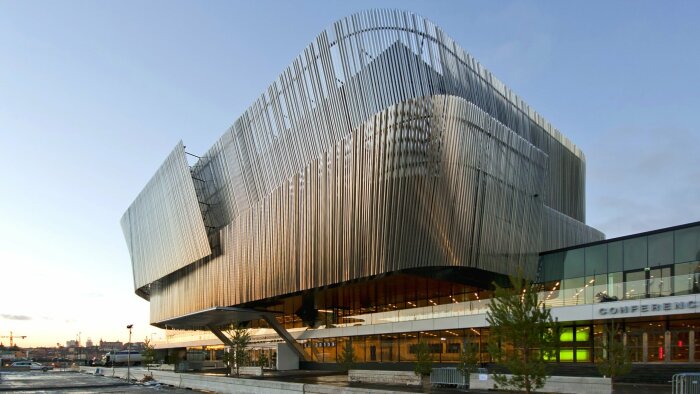 Modernt konferenscenter med böljande fasad av metallspjälor och stora glaspartier, omgiven av stadsmiljö och parkering, under klar himmel.