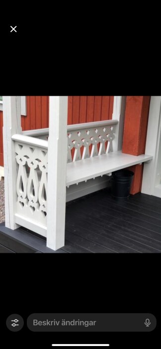 Vit, dekorerad träbänk vid röd vägg, placerad på en svart veranda. Svart kruka syns under bänken.