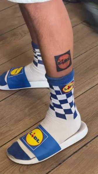 Ben med blåvita Lidle-strumpor och sandaler samt en tatuering på vaden med ett rött märke, mot en träparkett.